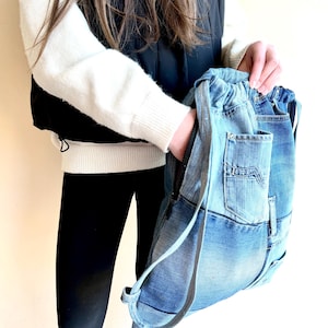 Op de afbeelding: Een denim rugzak met een trekkoordsluiting en een ritsvak. De tas is gemaakt van gerecyclede blauwe jeans en heeft de originele zakken en naden. De bandjes zijn lichtgrijs.