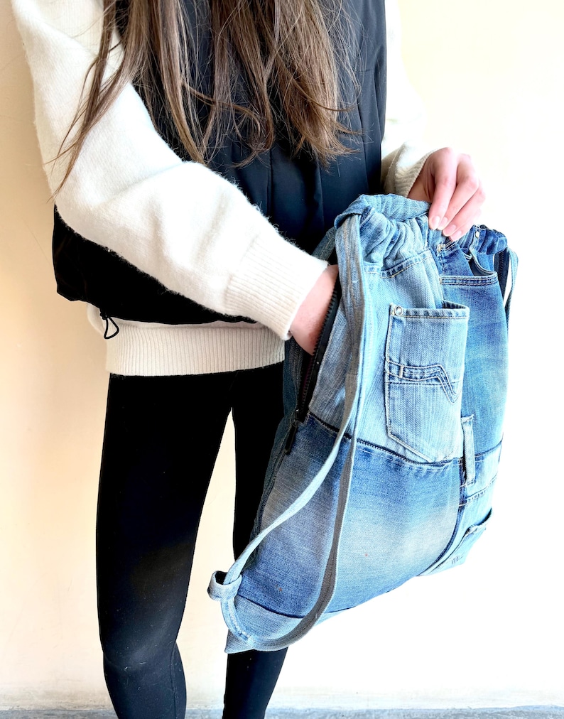 Op de afbeelding: Een denim rugzak met een trekkoordsluiting en een ritsvak. De tas is gemaakt van gerecyclede blauwe jeans en heeft de originele zakken en naden. De bandjes zijn lichtgrijs.