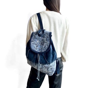 Può includere: Uno zaino realizzato in tessuto denim blu scuro con una patta e una sezione inferiore in tessuto a motivo grigio e blu screziato. La borsa include una chiusura a coulisse con cordini chiari e spallacci regolabili in denim scuro.