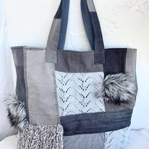 Könnte beinhalten: Eine Patchwork-Tragetasche mit verschiedenen Stoffbahnen in Grau- und Anthrazittönen. Das Design umfasst Abschnitte aus strukturiertem Strick, glattem Denim und zotteligem Stoff, akzentuiert durch zwei flauschige Kunstfell-Bommeln an den Seiten.