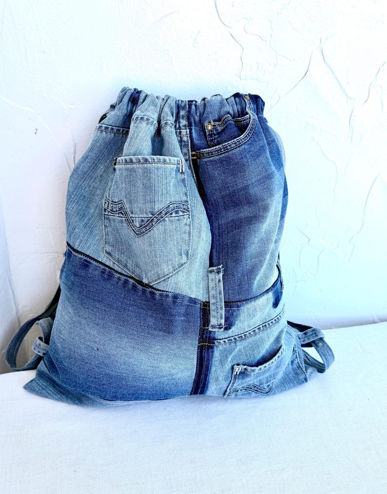 Op de afbeelding: Een rugzak met trekkoord gemaakt van gerecyclede spijkerbroeken. De tas heeft verschillende tinten blauwe denim, met zichtbare zakken en naden. Hij heeft schouderbanden en een trekkoordsluiting.