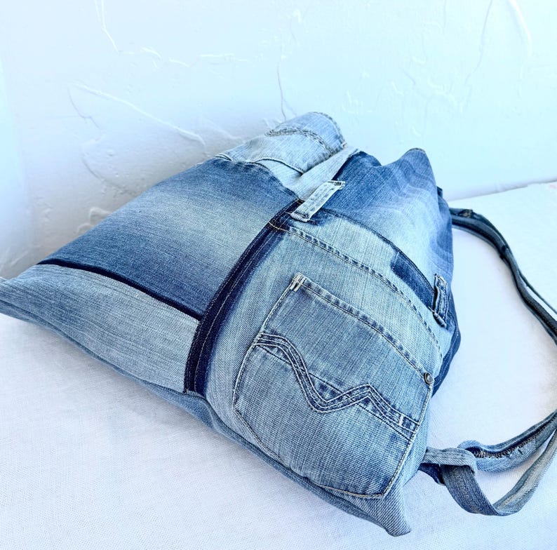 Op de afbeelding: Een denim tote bag gemaakt van gerecyclede jeans. De tas heeft een patchwork ontwerp met verschillende tinten blauw denim, inclusief zakken en naden. De bandjes zijn gemaakt van hetzelfde denim materiaal.