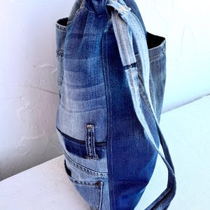 Op de afbeelding: Een tote bag met trekkoord gemaakt van hergebruikte spijkerbroeken. De tas heeft verschillende tinten blauwe denim, met zichtbare zakken en naden. De bandjes zijn gemaakt van hetzelfde denim materiaal.
