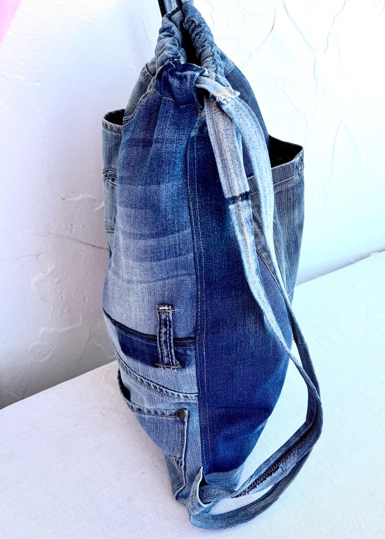 Op de afbeelding: Een tote bag met trekkoord gemaakt van hergebruikte spijkerbroeken. De tas heeft verschillende tinten blauwe denim, met zichtbare zakken en naden. De bandjes zijn gemaakt van hetzelfde denim materiaal.