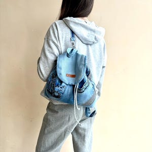 Op de afbeelding: Lichtblauwe denim rugzak met een klepsluiting en wit trekkoord. De rugzak heeft een patchwork ontwerp met verschillende denim wassingen en een klein bruin label. De bandjes zijn blauw.