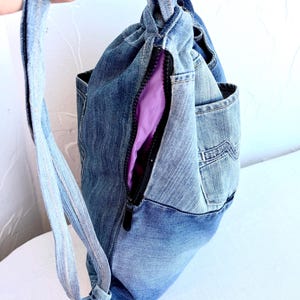 Op de afbeelding: Een denim rugzak met een trekkoordsluiting en een zijrits. De tas is gemaakt van hergebruikte spijkerbroeken en heeft een paarse binnenvoering. De riem is gemaakt van hetzelfde denim materiaal.