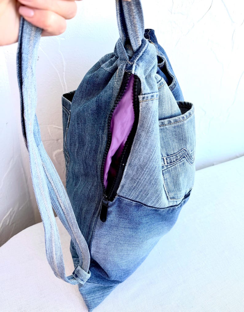 Op de afbeelding: Een denim rugzak met een trekkoordsluiting en een zijrits. De tas is gemaakt van hergebruikte spijkerbroeken en heeft een paarse binnenvoering. De riem is gemaakt van hetzelfde denim materiaal.