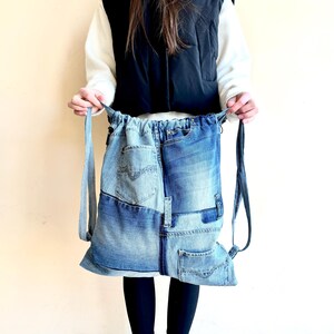 Op de afbeelding: Een denim rugzak gemaakt van hergebruikte jeans. De rugzak heeft een patchwork ontwerp met verschillende tinten blauwe denim en zichtbare zakken. Het heeft verstelbare bandjes en een trekkoordsluiting. De tas wordt vastgehouden door een persoon die een zwart vest en zwarte leggings draagt.