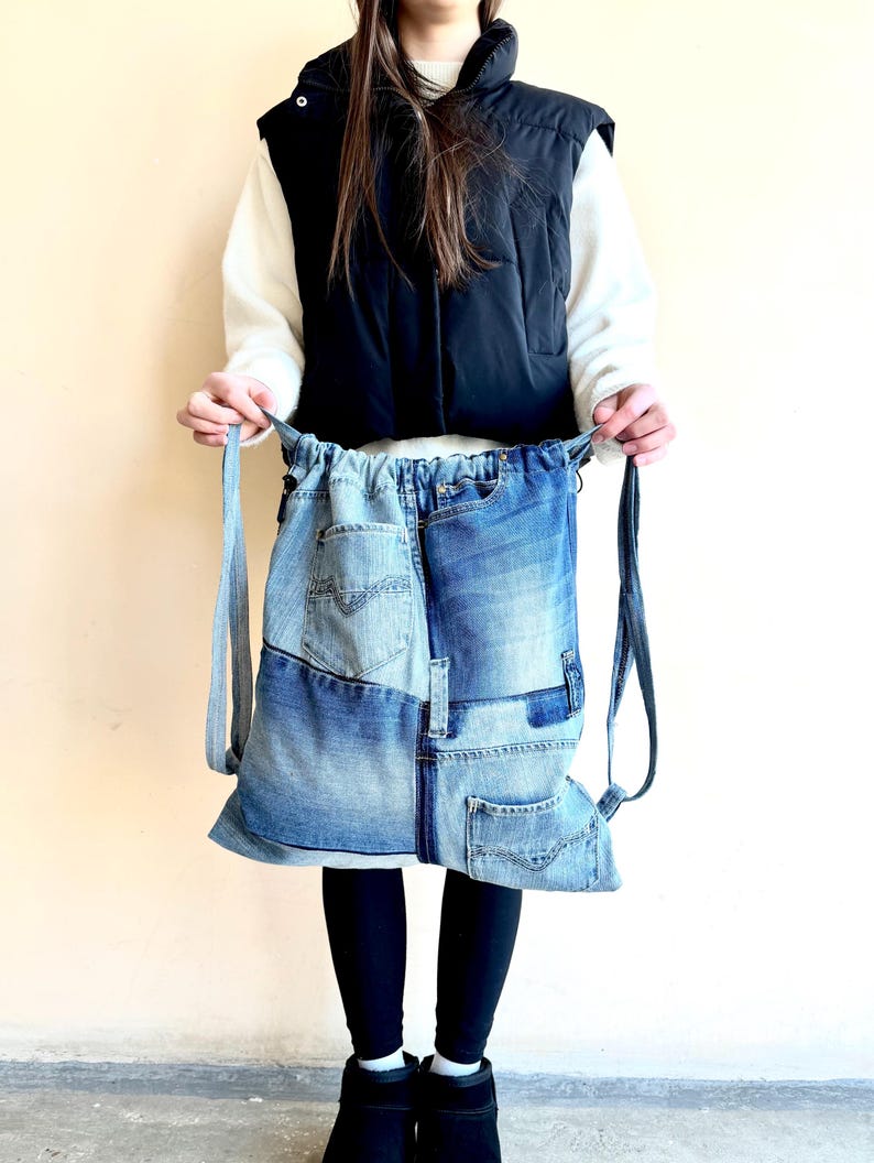 Op de afbeelding: Een denim rugzak gemaakt van hergebruikte jeans. De rugzak heeft een patchwork ontwerp met verschillende tinten blauwe denim en zichtbare zakken. Het heeft verstelbare bandjes en een trekkoordsluiting. De tas wordt vastgehouden door een persoon die een zwart vest en zwarte leggings draagt.