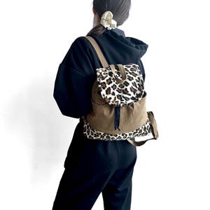 Puede incluir: Una mochila con cuerpo de lona color canela y una solapa y base de piel sintética con estampado de leopardo. La bolsa incluye correas de hombro ajustables de lona color canela, un asa superior y un cierre de cordón negro en la parte central.