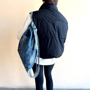 Op de afbeelding: Een denim rugzak met trekkoord, een voorvak en lichtblauwe bandjes wordt over de schouder gedragen. De tas is gemaakt van gerecyclede jeans. De persoon draagt een zwart puffer vest, een witte trui en zwarte leggings.