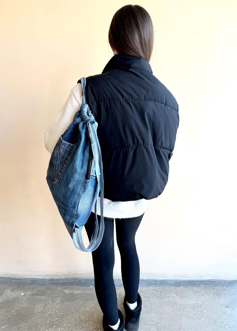 Op de afbeelding: Een denim rugzak met trekkoord, een voorvak en lichtblauwe bandjes wordt over de schouder gedragen. De tas is gemaakt van gerecyclede jeans. De persoon draagt een zwart puffer vest, een witte trui en zwarte leggings.