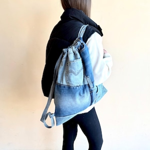 Op de afbeelding: Een lichtblauwe denim rugzak met trekkoord. De rugzak heeft een voorvak en verstelbare bandjes. De persoon op de foto draagt een zwarte puffer vest en zwarte leggings.