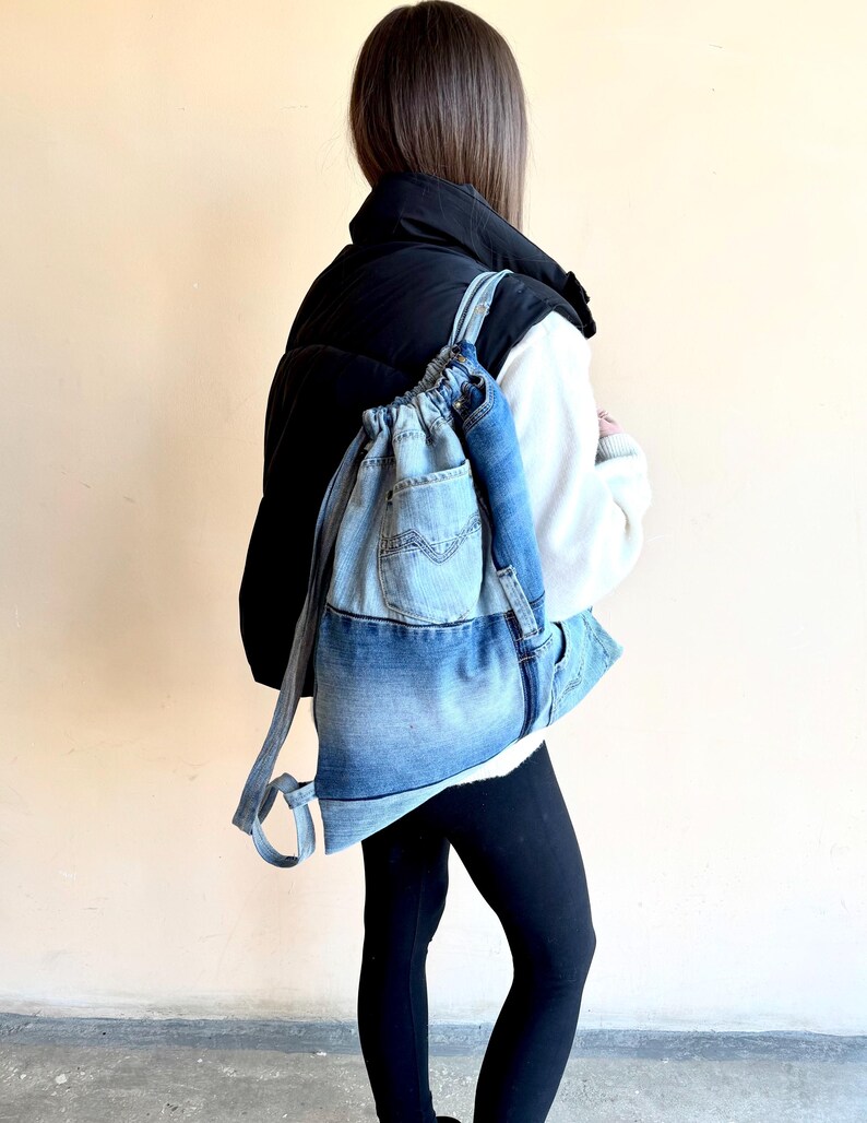 Op de afbeelding: Een lichtblauwe denim rugzak met trekkoord. De rugzak heeft een voorvak en verstelbare bandjes. De persoon op de foto draagt een zwarte puffer vest en zwarte leggings.