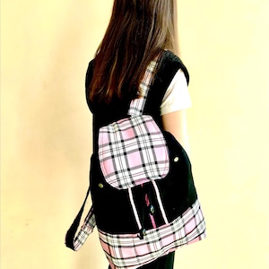 May include: Black backpack with pink and white plaid accents. The backpack has a flap pocket and adjustable straps. The bag is worn over a white t-shirt and black vest.