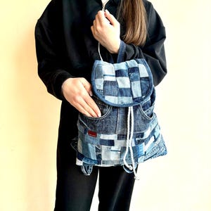 Può includere: Uno zaino in denim blu realizzato con jeans riciclati. Lo zaino ha una chiusura con coulisse e una patta con una tasca.