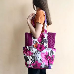 May include: A large floral tote bag with a purple lining and two side pockets. The bag is made of a white fabric with a pink, purple, and orange floral print.