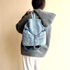 Op de afbeelding: Een lichtblauwe denim rugzak met een trekkoordsluiting en voorzakken. De rugzak heeft blauwe bandjes en een rits. De tas wordt gedragen over een grijze hoodie.