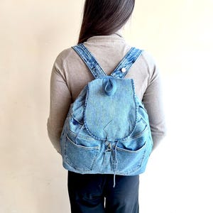 Pode incluir: Uma mochila de ganga azul de lavagem média com uma aba dobrável com um laço central, dois bolsos frontais com costuras visíveis e alças de ombro ajustáveis em ganga. O saco é feito de tecido de ganga reutilizado com uma textura azul clássica.