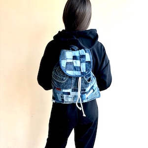 May include: A blue denim backpack made from recycled jeans. The backpack has a drawstring closure and a flap with a buckle.