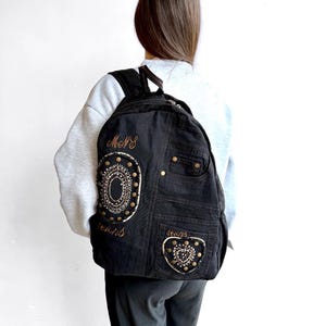 May include: A black denim backpack featuring a large circular patch and a smaller heart-shaped patch, both embellished with metallic studs and intricate embroidery. The text "MNS" and "jeans" is embroidered on the front, alongside a functional pocket.