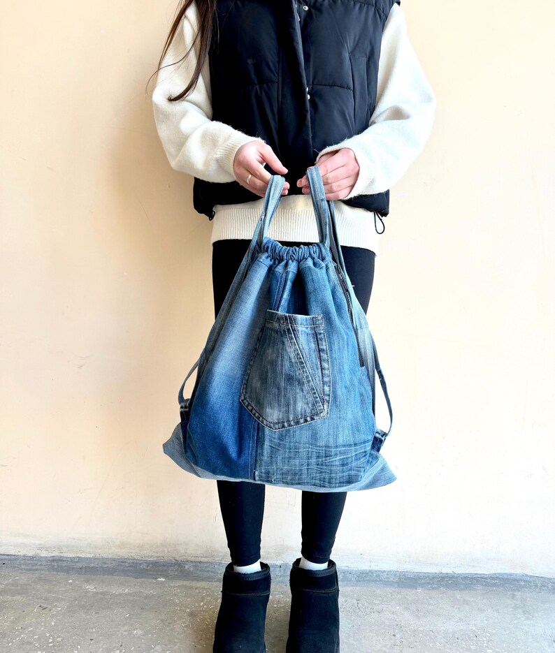 Op de afbeelding: Een blauwe denim rugzak met trekkoord en een voorvak. De tas is gemaakt van gerecycled denim en heeft verstelbare bandjes. De tas wordt vastgehouden door een persoon die een zwart vest, witte trui, zwarte legging en zwarte laarzen draagt.