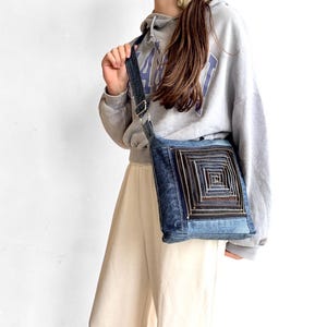 May include: A crossbody bag made from blue denim fabric featuring a square-shaped decorative panel on the front. The panel is constructed from multiple concentric layers of dark brown and black zips arranged in a repeating square pattern.