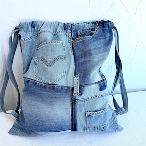 Op de afbeelding: Een rugzak met trekkoord gemaakt van hergebruikte spijkerbroeken. De tas heeft een patchwork-ontwerp met verschillende tinten blauwe denim, inclusief zakken en naden van de originele jeans. De bandjes zijn gemaakt van hetzelfde denim materiaal.