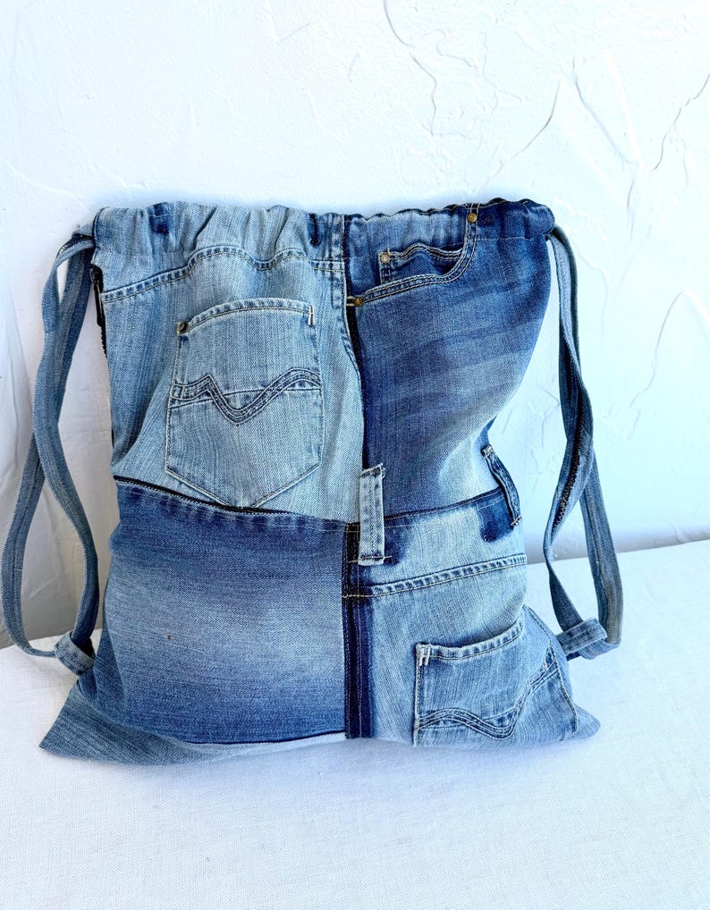 Op de afbeelding: Een rugzak met trekkoord gemaakt van hergebruikte spijkerbroeken. De tas heeft een patchwork-ontwerp met verschillende tinten blauwe denim, inclusief zakken en naden van de originele jeans. De bandjes zijn gemaakt van hetzelfde denim materiaal.