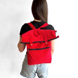 May include: A bright red corduroy backpack featuring a fold-over top design with a black zipper closure, two shoulder straps, a top carry handle, and an additional horizontal zippered pocket on the front section of the bag.
