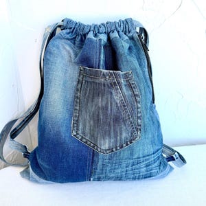 Op de afbeelding: Een blauwe denim rugzak met trekkoord en een donkerblauwe zak. De rugzak heeft zwarte bandjes en een zwarte rits. Het denim heeft verschillende blauwtinten, waardoor een patchwork-effect ontstaat. De tas is ontworpen om spullen te dragen.