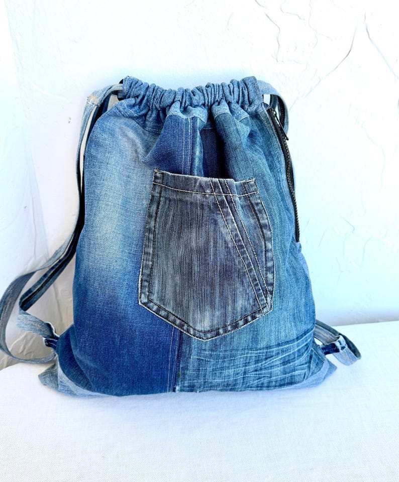 Op de afbeelding: Een blauwe denim rugzak met trekkoord en een donkerblauwe zak. De rugzak heeft zwarte bandjes en een zwarte rits. Het denim heeft verschillende blauwtinten, waardoor een patchwork-effect ontstaat. De tas is ontworpen om spullen te dragen.