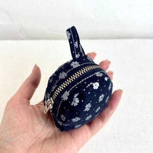 May include: A small, round, dark blue fabric pouch featuring a repeating white floral pattern. The pouch includes a metallic zip closure that runs across the top and a matching fabric loop handle attached to the upper edge for easy carrying.