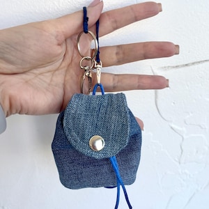 May include: A miniature denim backpack keyring with a silver button closure and blue drawstring. The keyring has a blue cord loop and a silver clasp and key ring.