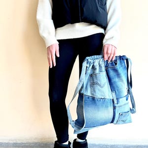 Op de afbeelding: Een denim rugzak met een trekkoordsluiting. De tas is gemaakt van hergebruikte blauwe jeans, met zakken en naden. De bandjes zijn ook gemaakt van denim. De tas wordt vastgehouden door een persoon die een zwarte legging, zwarte laarzen, een witte trui en een zwart vest draagt.