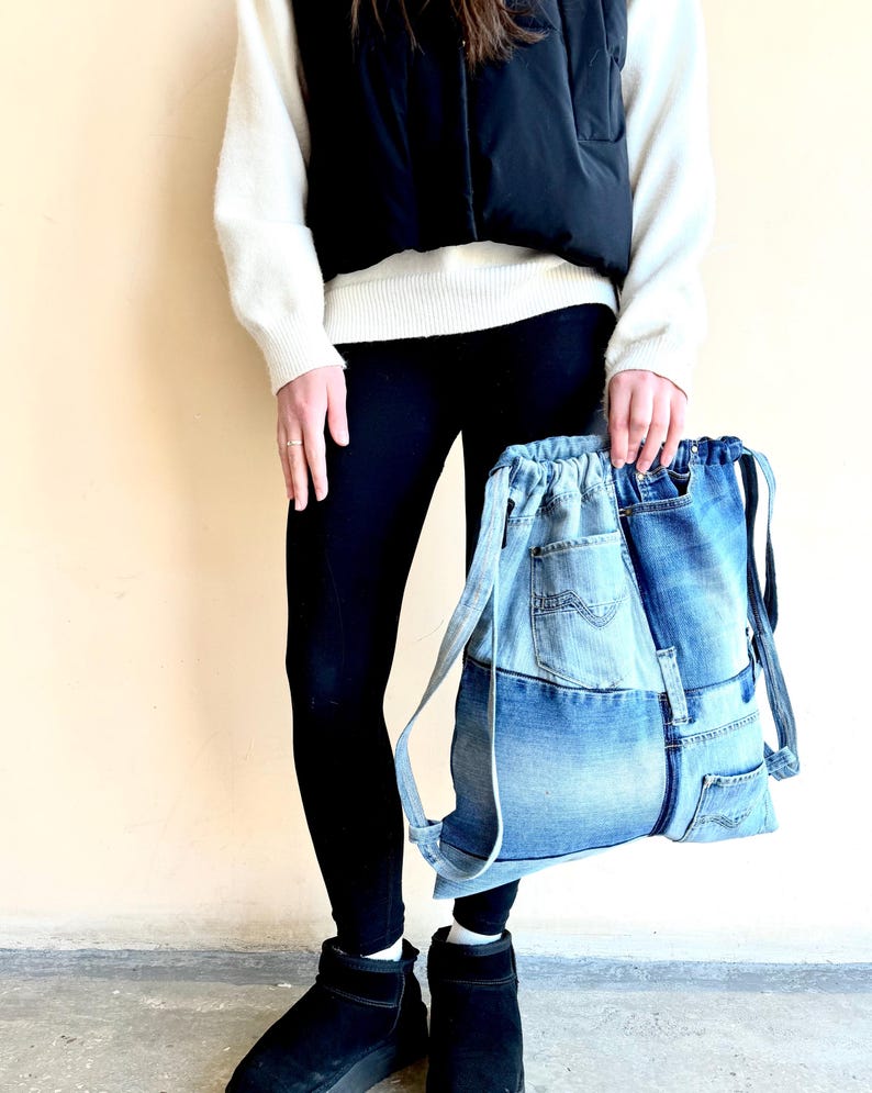 Op de afbeelding: Een denim rugzak met een trekkoordsluiting. De tas is gemaakt van hergebruikte blauwe jeans, met zakken en naden. De bandjes zijn ook gemaakt van denim. De tas wordt vastgehouden door een persoon die een zwarte legging, zwarte laarzen, een witte trui en een zwart vest draagt.