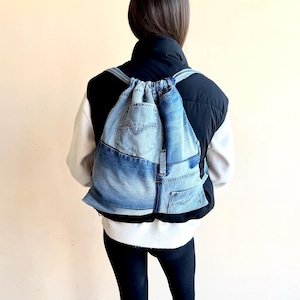 May include: A denim drawstring backpack made from repurposed jeans. The backpack features a patchwork design with various shades of blue denim, a front pocket, and adjustable straps. The bag is worn over a black and white outfit.