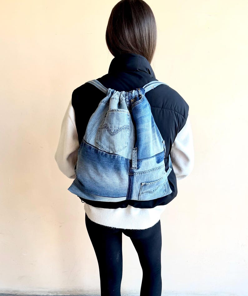 Op de afbeelding: Een denim rugzak met trekkoord gemaakt van gerecyclede jeans. De rugzak heeft een patchwork ontwerp met verschillende tinten blauwe denim, een voorvak en verstelbare bandjes. De tas wordt gedragen over een zwart-witte outfit.