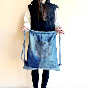 Op de afbeelding: Blauwe denim rugzak met trekkoord en voorvak. De rugzak is gemaakt van gerecycled denim en heeft een patchwork design met verschillende blauwtinten. De bandjes zijn ook van denim.