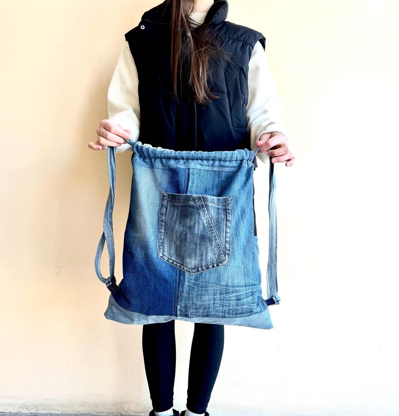 Op de afbeelding: Blauwe denim rugzak met trekkoord en voorvak. De rugzak is gemaakt van gerecycled denim en heeft een patchwork design met verschillende blauwtinten. De bandjes zijn ook van denim.