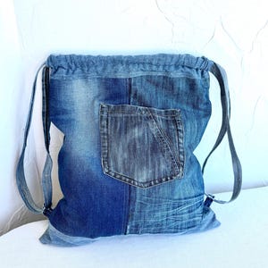 Op de afbeelding: Blauwe denim rugzak met trekkoord en voorvak. De rugzak is gemaakt van gerecycled denim en heeft verstelbare bandjes. Het denim heeft verschillende blauwtinten en een versleten uitstraling.