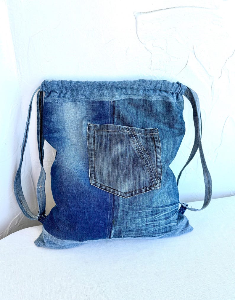 Op de afbeelding: Blauwe denim rugzak met trekkoord en voorvak. De rugzak is gemaakt van gerecycled denim en heeft verstelbare bandjes. Het denim heeft verschillende blauwtinten en een versleten uitstraling.