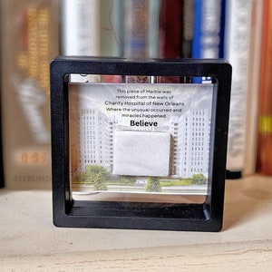 May include: A black framed display case with a white marble piece inside. The case has a white background with text that reads: "This piece of Marble was removed from the walls of Charity Hospital of New Orleans Where the unusual occurred and miracles happened. Believe."