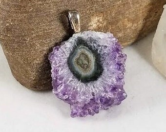 May include: A solar amethyst pendant with a silver bail. The pendant features a central dark green and gray pattern surrounded by a ring of white and a border of light purple crystals. The pendant is a unique piece of jewelry.