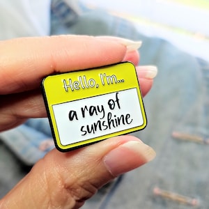 May include: A rectangular enamel pin with a yellow background and black border. The pin reads "Hello, I'm..." at the top and "a ray of sunshine" in a white rectangle below. The pin is held by a person.