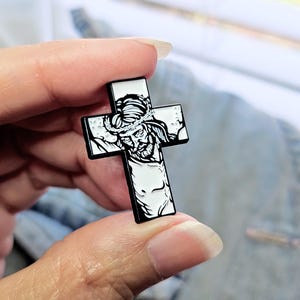 May include: A black and white enamel pin featuring a depiction of Jesus Christ on a cross. The pin is a religious symbol and is perfect for adding a touch of faith to your clothing or bag.