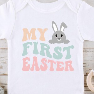 May include: White baby onesie with the text "MY FIRST EASTER" in pastel colours. A grey bunny rabbit is peeking out from behind the text.