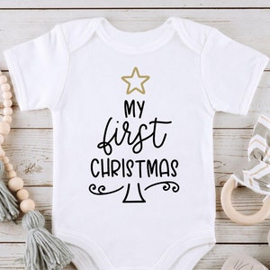 以下が含まれることがあります： 白いベビーロンパースに、黒と金のデザインで「初めてのクリスマス」と書かれており、スタイリッシュなクリスマスツリーのグラフィックが描かれています。