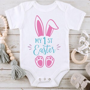 May include: White baby onesie with pink bunny ears and pink footprints. The onesie has the text "My 1st Easter" in blue.