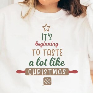 May include: White sweatshirt with the text "It's beginning to taste a lot like Christmas" in a Christmas tree design. The design includes a star, rolling pin, and cookie graphic. The sweatshirt is a digital download.